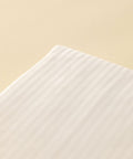 Detailaufnahme der Ecke eines Kissenbezugs aus weißer Microstripe Bettwäsche mit dezent glänzenden, eingewebten Längsstreifen vor einem warmen, beigen Hintergrund.
