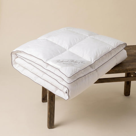 Excellence Eiderdaunendecke sauber gefaltet auf einem Hocker, präsentiert mit hochwertiger Verarbeitung und luxuriöser Daunenfüllung.