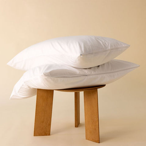 Zwei weiße Kissenschonbezüge dekorativ auf einem Holzhocker präsentiert, mit pflegeleichtem Stoff und schlichtem Design